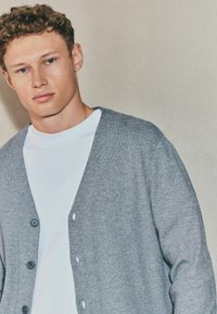 Tânăr cu păr creț de culoare castanie deschisă, purtând o cămașă albă și un cardigan gri cu nasturi, stând în fața unui fundal simplu și deschis.