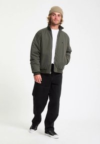 Olive green bomber jacket with a high collar, black pants, and black sneakers. The jacket features a zipper and a relaxed fit design.