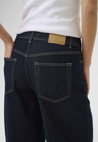 Jeans de mezclilla azul oscuro con costuras doradas en contraste. Presenta dos bolsillos traseros y una etiqueta de cuero marrón en la cinturilla.