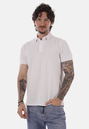 Uomo con tatuaggi su entrambe le braccia, indossa una polo bianca a maniche corte e jeans blu, in piedi davanti a uno sfondo bianco uniforme.