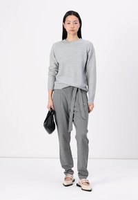 Grau gestrickter Pullover mit geripptem Saum, kombiniert mit grauen, taillierten Hosen, die an der Taille gebunden sind. Eine schwarze Clutch und flache Schuhe runden den Look ab.