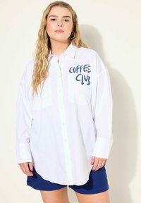 Camisa blanca de manga larga con botones, dos bolsillos en el pecho y el texto "COFFEE CLUB" en color azul marino. El modelo la combina con pantalones cortos azules. Textura de tela suave.