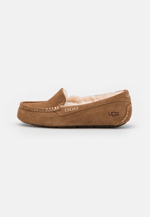 Chausson mocassin en daim marron avec doublure en laine beige et logo UGG sur le talon, présentant des coutures décoratives et une semelle en caoutchouc.