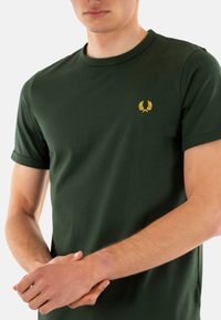 Camiseta de algodón de color verde oscuro con mangas cortas, que presenta un pequeño emblema bordado amarillo en el pecho. Textura suave, diseño de cuello redondo.