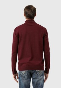 Burgunderfarbener Strickpullover mit hohem Kragen, gerippten Bündchen und Saum, kombiniert mit blauen Jeans mit dekorativen Gesäßtaschen.