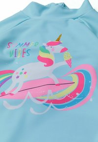 Un t-shirt de surf bleu clair avec un dessin de licorne surfant sur une planche. Le design comprend des vagues colorées et le texte "Vibes d'été" dans des couleurs vibrantes.