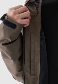 Main ajustant la fermeture éclair sur la manche d'une veste marron avec une fermeture à crochet et bande auto-agrippante noire, montrant la doublure de la veste et les vêtements intérieurs noirs.