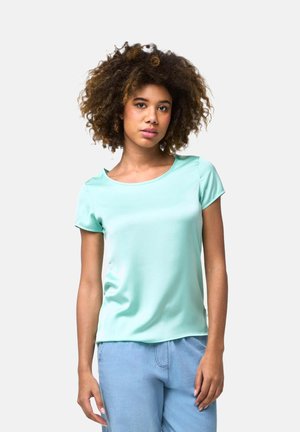 Jeune femme aux cheveux bouclés portant un haut vert clair à manches courtes et un pantalon bleu clair, debout devant un fond blanc uni.