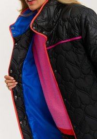 Prošívaná čierna bunda s ružovou podšívkou a modrou tkaninou, s oranžovými detailmi a zapínaním na patentky. Viditeľný textúrovaný vzor.
