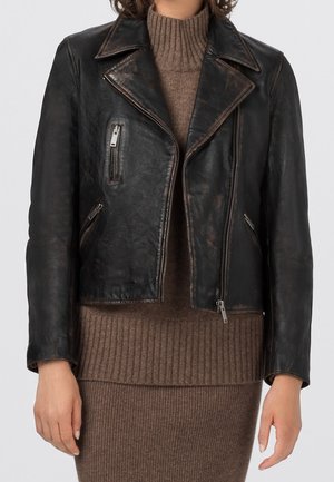 Osoba ubrana w czarną, postarzaną skórzaną kurtkę motocyklową na brązowym prążkowanym golfie i spódnicy.