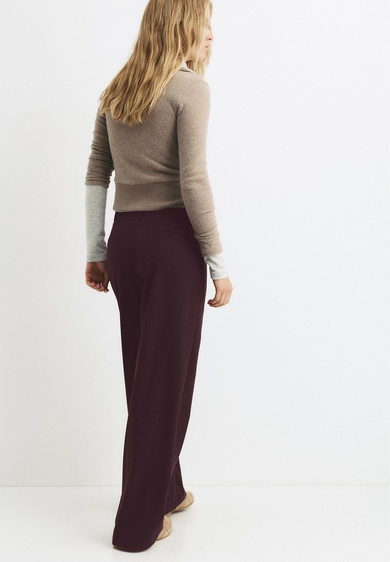 Femme aux longs cheveux blonds portant un haut à manches longues superposé beige et gris, ainsi qu'un pantalon large foncé, debout devant un fond blanc.