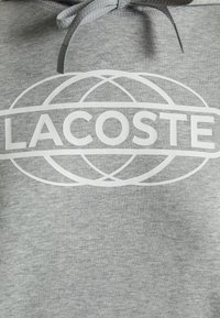 Sweat à capuche gris avec texte blanc de la marque Lacoste et motif globe superposé centré sur la poitrine sous une capuche ajustée à cordon.