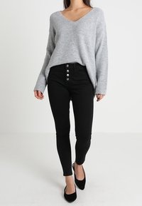 Grau gerippter Pullover mit V-Ausschnitt, kombiniert mit schwarzen High-Waist Skinny-Jeans mit fünf silbernen Knöpfen und strukturiertem Stoff.