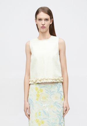 Woman with straight brown hair wears sleeveless cream top with ruffle hem and floral patterned pastel skirt against plain background.