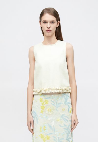 Femme aux cheveux bruns lisses portant un haut crème sans manches avec ourlet volanté et une jupe pastel à motif floral sur un fond uni.