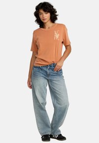 Perzikkleurig T-shirt met korte mouwen en een grafisch ontwerp op de borst, gecombineerd met lichtblauwe wijde jeans en zwarte sneakers.