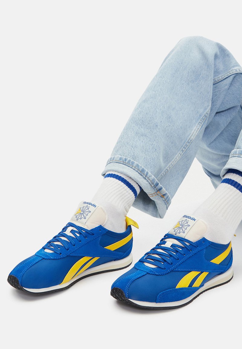 Niebiesko-żółte buty sportowe z gładką syntetyczną cholewką, białą gumową podeszwą i logo na płaskim języku. Noszone z jasnymi dżinsami.