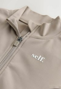 Light beige zip-up jacket with a high collar, featuring a smooth texture, silver-tone zipper, and a small, white logo on the front.