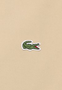 Zelená krokodýlí logo vyšívané na béžovém textilním pozadí s bílým obrysem, obsahující detailní šití a viditelný úsměv.
