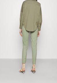Camicia verde oliva, comoda e a maniche lunghe, abbinata a pantaloni verdi chiari a sigaretta e sandali beige con lacci. Vista posteriore.
