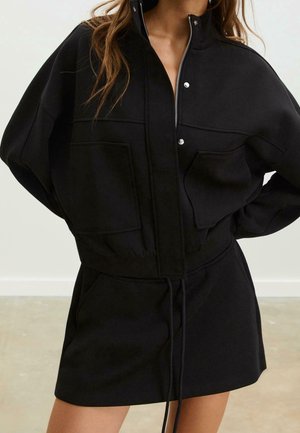 Femme portant une veste noire zippée avec boutons-pression et grandes poches avant, associée à une jupe noire assortie avec taille à cordon.