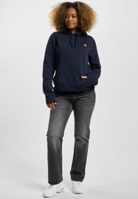 Marineblå hettegenser laget av mykt stoff med kengurulomme, med en logo på brystet. Kombinert med grå rette jeans og hvite sneakers.
