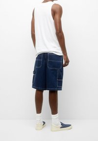 Pantalones cortos de mezclilla tipo cargo con bolsillos profundos, color azul oscuro y costuras en contraste, combinados con una camiseta sin mangas blanca y zapatillas claras.