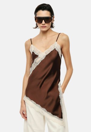 Donna che indossa una camicia di raso marrone con bordo in pizzo bianco, grandi occhiali da sole scuri e pantaloni bianchi, in piedi con una mano in tasca.