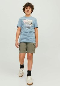 T-shirt in cotone azzurro chiaro con stampa grafica, abbinata a pantaloni cargo verde oliva. Sneakers con lacci bianchi e calzini neri alla caviglia.