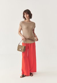 Top beige lavorato a maglia con maniche corte, abbinato a pantaloni rossi a gamba larga. In una mano è tenuta una piccola borsa in suede marrone con chiusura in argento.