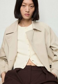 Chaqueta beige ajustada sobre un cárdigan de canalé crema con detalles de botones, combinada con pantalones marrones de talle alto. Textura suave.