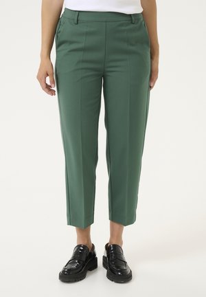 Grüne maßgeschneiderte knöchellange Hosen, kombiniert mit schwarzen klobigen Loafers und einem weißen, in die Hose gesteckten Oberteil, gezeigt an einer stehenden Person.