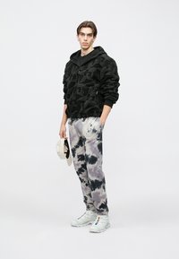 Sort teksturert hettejakke med glidelås, grå tie-dye joggebukser og hvite joggesko. Modellen holder en beige caps og viser frem et avslappet antrekk.