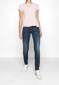 Nainen, jolla on päällään vaaleanpunainen Abercrombie & Fitch t-paita, tummansiniset skinny-farkut ja valkoiset lenkkarit, seisoo yksinkertaista valkoista taustaa vasten.