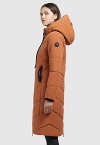 Paddad orange kappa med huva, featuring en svart dragkedja, rund ficka och chevronquiltat mönster. Den texturerade tyget ger isolering.