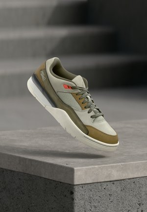 Sneaker marrone chiaro e beige con lacci grigi e dettaglio rosso che fluttua sopra una sporgenza di cemento con sfondo di scale sfocate.