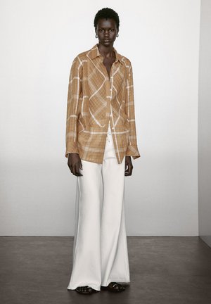 Chemise à manches longues à carreaux marron avec des lignes blanches, devant à boutons, coupe décontractée, associée à un pantalon large blanc et des sandales noires.