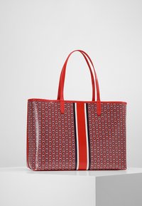 Czerwona torba tote z geometrycznym wzorem, z dwoma uchwytami i centralnym białym oraz ciemnoniebieskim paskiem. Gładka faktura zewnętrzna.