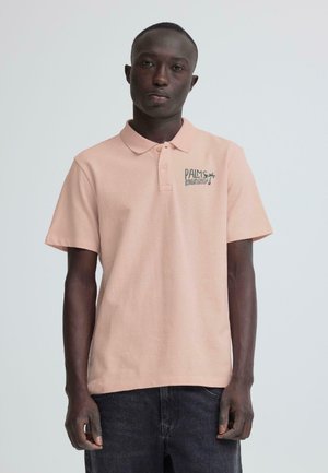 Vīrietis valkā gaiši rozā polo kreklu ar uzrakstu "Palm Island" un palmas koka logo uz krūtīm, tumsas bikses, stāvot pret baltu fonu.