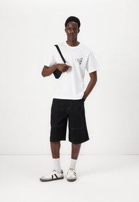 Vit t-shirt med grafisk tryck, svarta shorts, vita sneakers med gråa och svarta accenter, samt en liten svart axelväska.