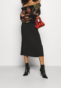 Donna che indossa un blusa nera floreale off-shoulder, una gonna midi nera, stivali alla caviglia neri con tacco, e tiene in mano una piccola borsa a tracolla rossa intrecciata.