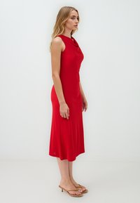 Rode, mouwloze midi-jurk met een cowl-halslijn. De stof is soepel en licht rekbaar, en eindigt net onder de knie. Gemaakt in combinatie met beige hoge sandalen.