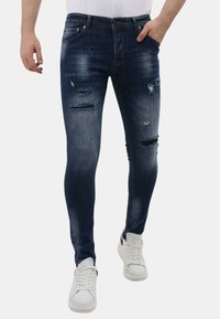 Slanke blauwe spijkerbroek met versleten plekken, vijf zakken en een middelhoge taille. Draag met witte sneakers voor een casual look.