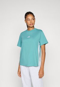 Turkos tränings t-shirt med korta ärmar, rundad halsringning och tre vita ränder längs sidorna. Har en liten märkeslogotyp på bröstet.