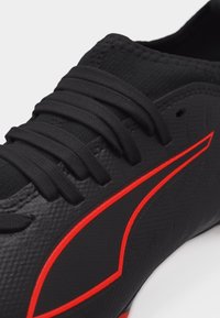 Κοντινό πλάνο μαύρου αθλητικού παπουτσιού με υφή στην επιφάνεια, μαύρα κορδόνια και έντονο κόκκινο γωνιώδες σχέδιο στη πλευρά.