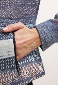 Veste à motifs multiples en tissu bleu texturé, avec une poche zippée et un col structuré souligné par des coutures apparentes.
