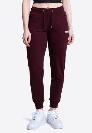 Femme portant un pantalon de jogging bordeaux avec un cordon de serrage et des baskets blanches, debout devant un fond uni.