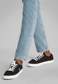 Asmuo dėvi šviesiai mėlynas džinsines kelnes ir juodus žemus sportbačius su baltais raišteliais ir baltais padais, vaikščiojantį pilkame fone.