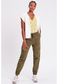 Cardigan senza maniche lavorato a maglia bianco, top con motivo geometrico giallo, pantaloni cargo verde oliva con polsini elasticizzati e scarpe da ginnastica di colore chiaro.