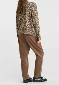 Femme portant un haut à manches longues imprimé léopard, un pantalon ample marron texturé et des chaussures noires à lacets, debout devant un fond uni.